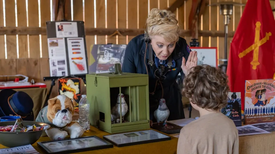 Woman with headset enthusiastically explains to a child at an educational display featuring pigeons, a plush dog, and historical posters. Warm, engaging atmosphere.