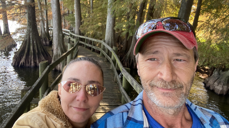 A man and woman posing on a wooden bridge through a forested area by a lake. They appear relaxed and happy, with fall foliage in the background.