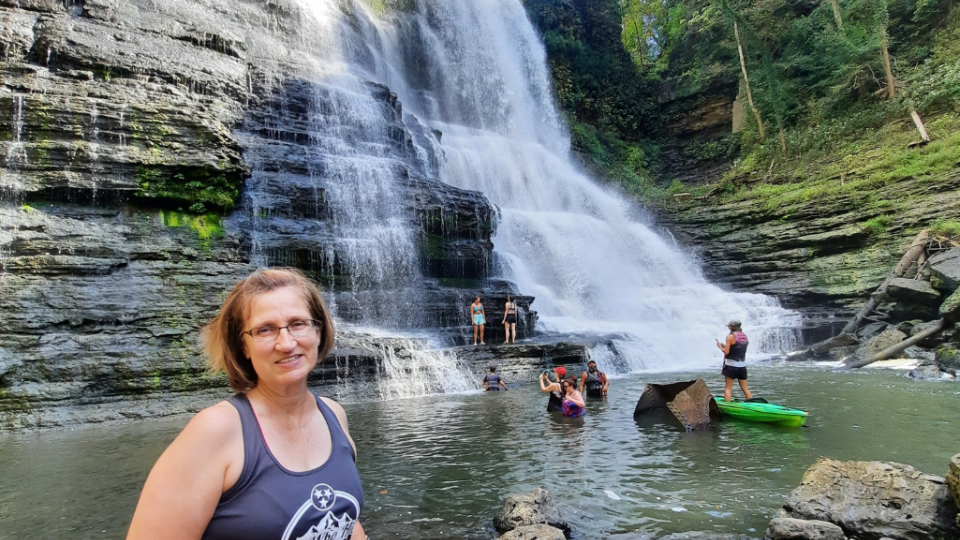 A smiling woman stands by a tranquil waterfall surrounded by lush greenery. People play in the water, creating a joyful, natural atmosphere.