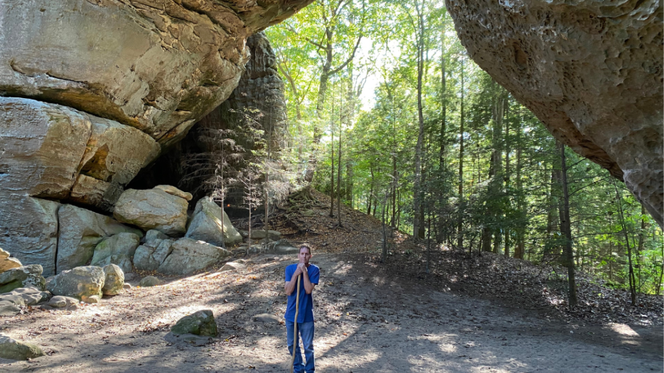 A person in a blue shirt stands under a large rock arch in a forest, with sunlight filtering through trees. The scene feels peaceful and awe-inspiring.