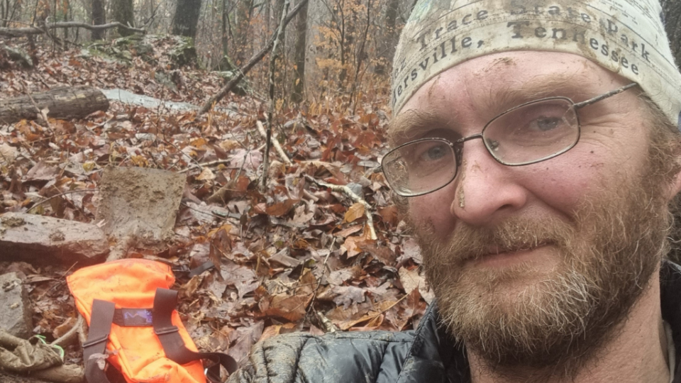 A bearded man wearing glasses and a bandana takes a selfie in a forest. He looks content. The background shows fallen leaves and an orange backpack.