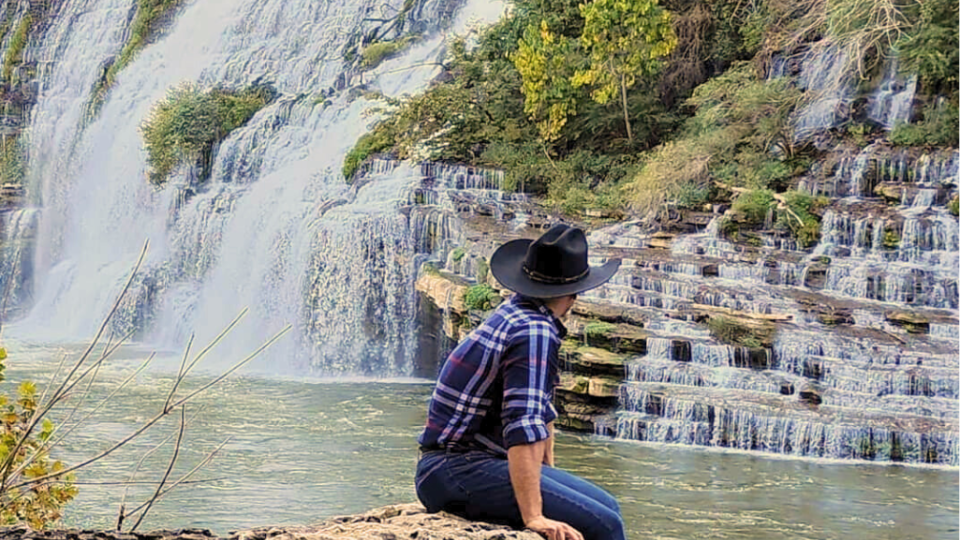A person in a plaid shirt and black hat sits on a rock, gazing at a cascading waterfall surrounded by lush greenery, creating a serene atmosphere.