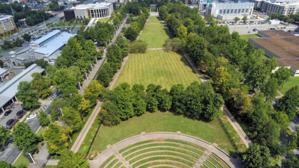 A drone shot captures the park’s central grassy lawn flanked by rows of trees, with the amphitheater in the foreground and city buildings visible in the distance.