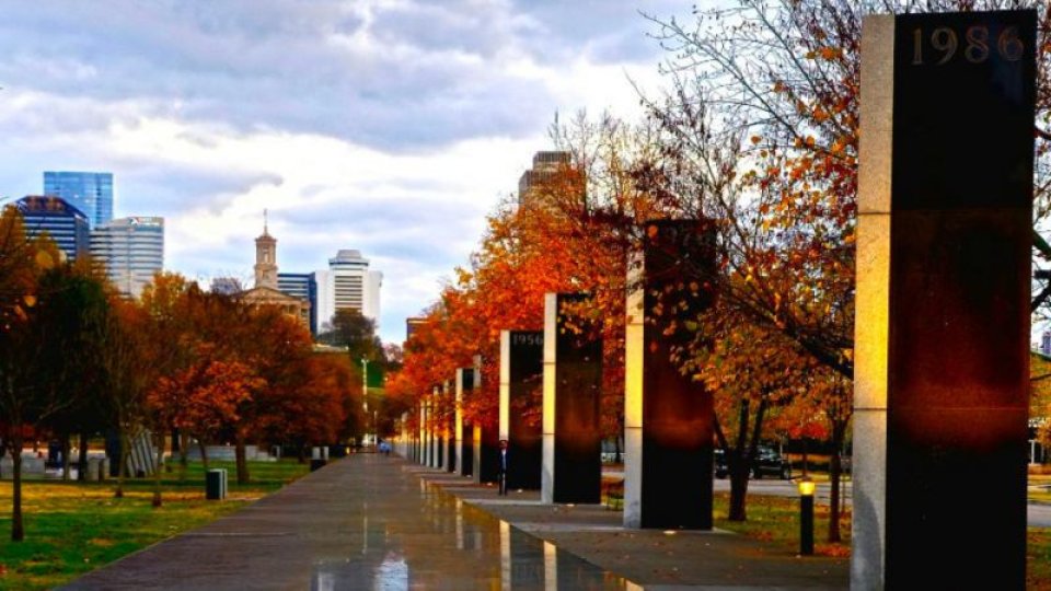 A row of black granite timeline markers lines a path through the park, each engraved with a year, set against colorful autumn foliage and a backdrop of the Nashville skyline.