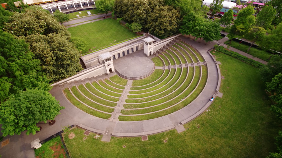 Aerial view of an outdoor amphitheater with semicircular steps and two towers. Surrounded by lush green trees and pathways, conveying tranquility and openness.