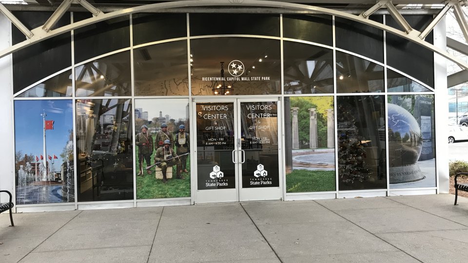 The front entrance of the Bicentennial Capitol Mall State Park Visitor Center features large glass panels decorated with scenic and historical images, including soldiers, a flag display, park columns, and a globe monument. The glass doors display operating hours and the Tennessee State Parks logo.