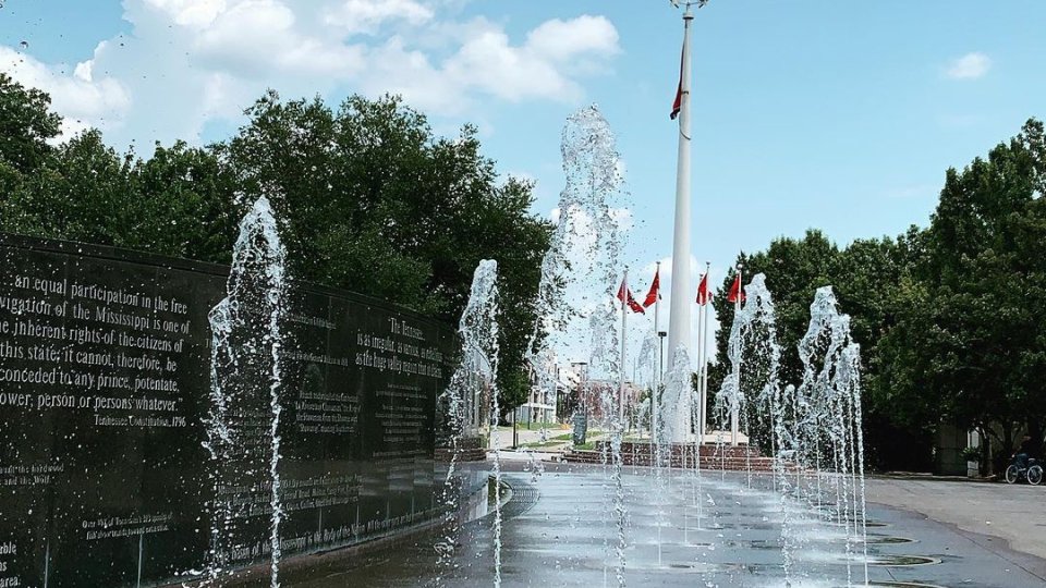 Fountains spray water upward in a plaza surrounded by green trees and a tall flagpole with flags. The sky is blue with fluffy clouds, creating a lively atmosphere.