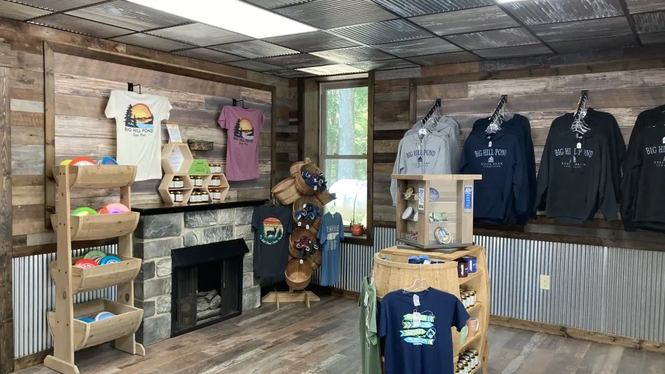 Wooden shop interior with rustic decor, featuring colorful disc golf discs on shelves, graphic T-shirts, and hoodies displayed on walls. Cozy and inviting.