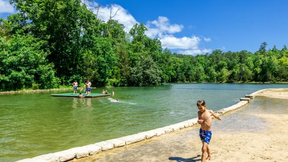Boy in swim trunks on sandy shore, others swimming in a lake with lush green trees.