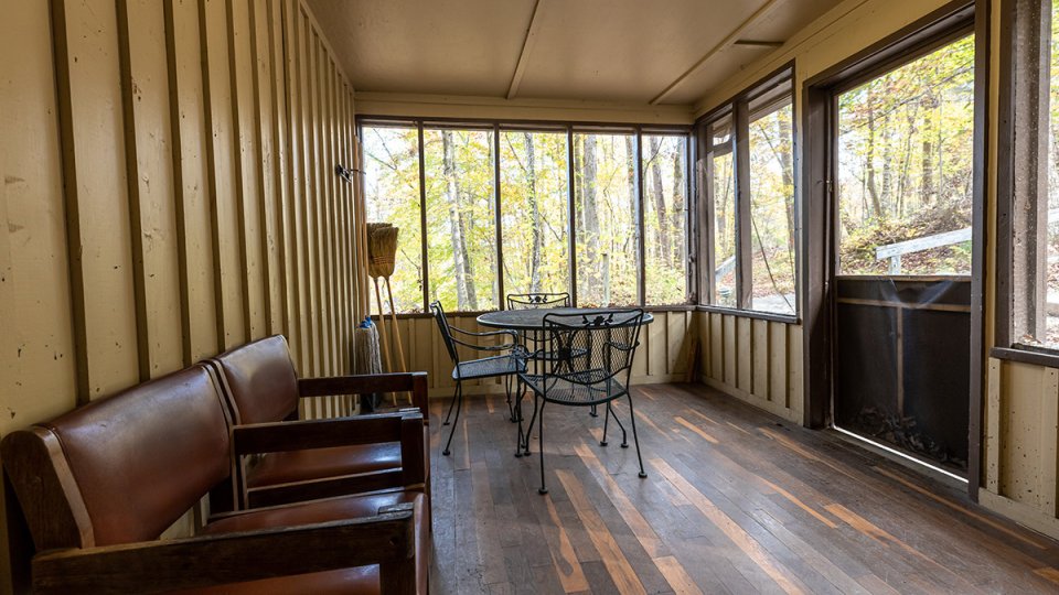 Breezeway inside a cabin on a sunny day with two chairs, a dinig room table and a screened in wall. 
