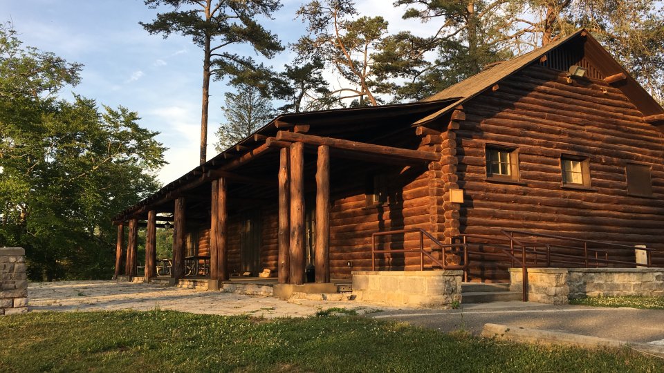 Log cabin beneath tall pine trees, bathed in warm sunlight. The rustic structure has a stone foundation and wooden porch, evoking a peaceful, natural ambiance.