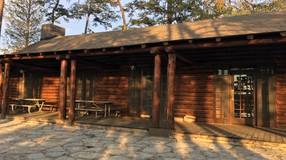A rustic log cabin with a wooden porch sits in front of a stone patio. Tall trees surround the cabin, casting long shadows in late afternoon sunlight.