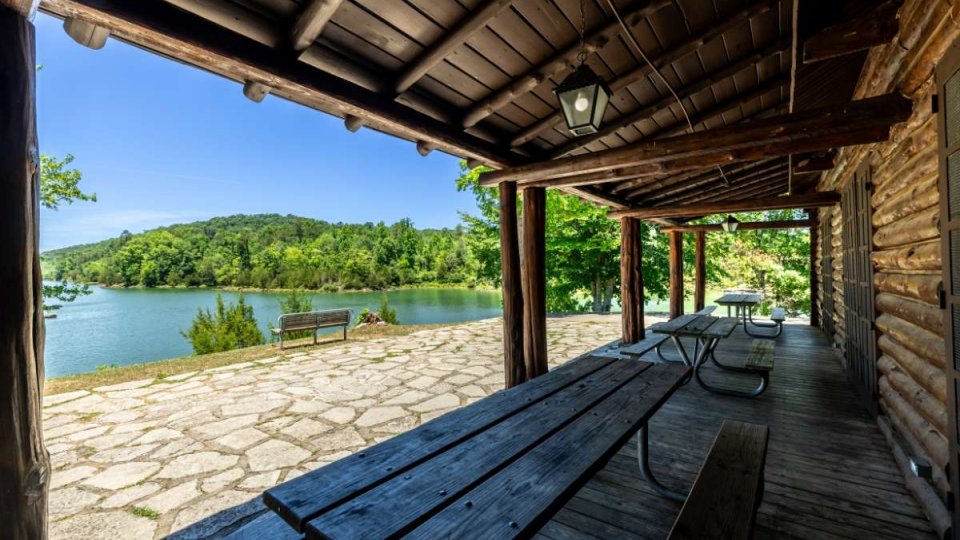 Wooden porch with picnic tables overlooking a serene lake and lush green hills under a blue sky. Peaceful and inviting outdoor scene.