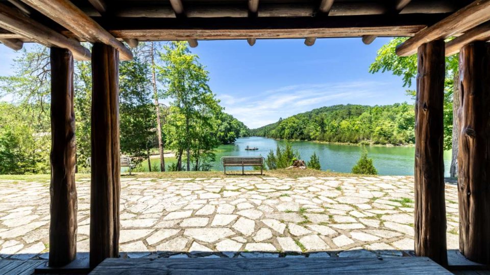 View from a wooden gazebo, overlooking a tranquil lake and lush green hills. A stone patio with a bench under a clear blue sky creates a peaceful setting.