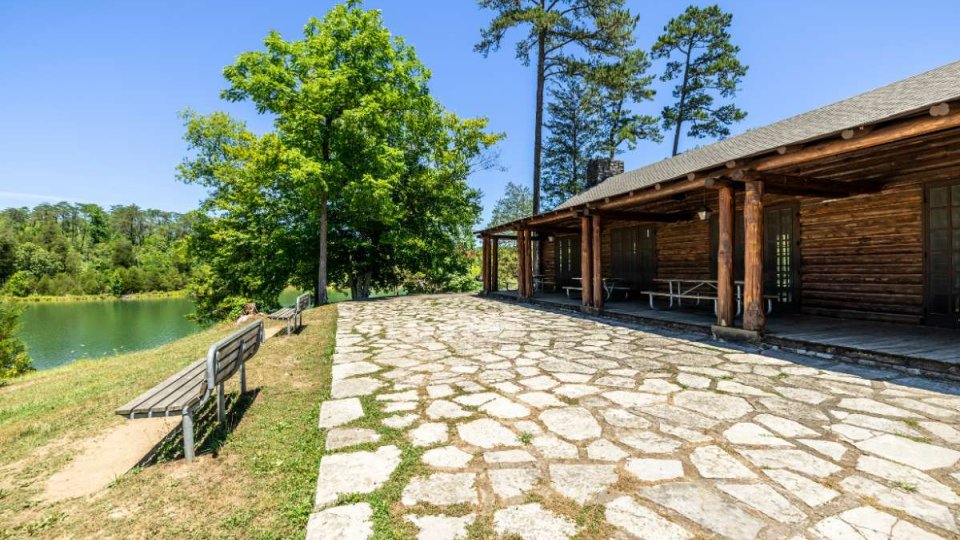 Log cabin with a stone path and wooden benches overlooks a serene lake. Tall trees and vibrant greenery create a peaceful, rustic atmosphere.