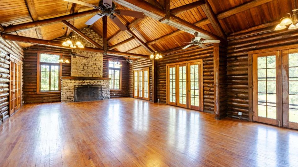Spacious log cabin interior with wooden beams, stone fireplace, and polished wooden floors. Bright natural light through multiple French doors. Cozy, rustic ambiance.