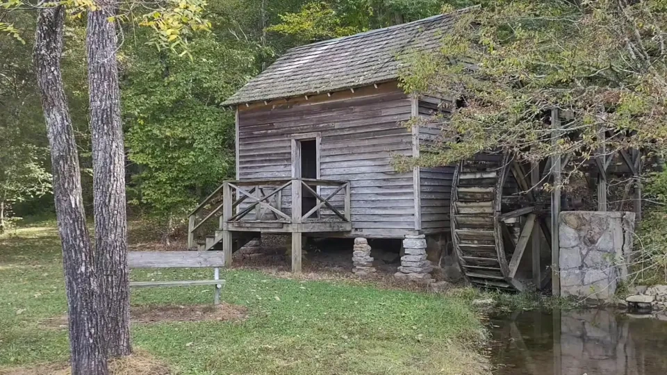 Old wooden watermill beside a serene stream in a lush, green forest. The structure is rustic, evoking a peaceful, timeless atmosphere.