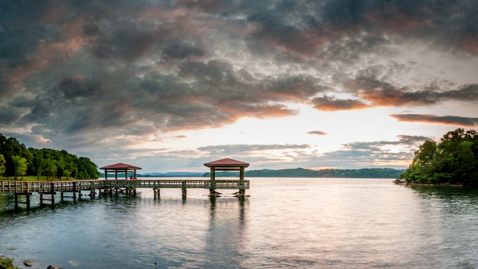 Sunset over a calm lake with wooden piers extending into the water. Dramatic clouds fill the sky, surrounded by lush green trees, creating a serene atmosphere.