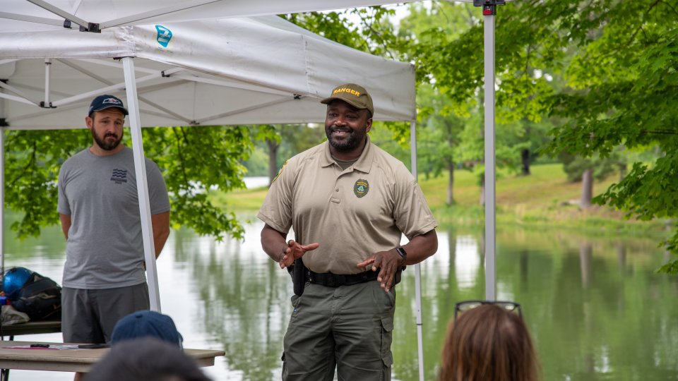 A park ranger speaks animatedly under a canopy near a pond, surrounded by green trees. Attendees listen attentively, creating a relaxed outdoor setting.