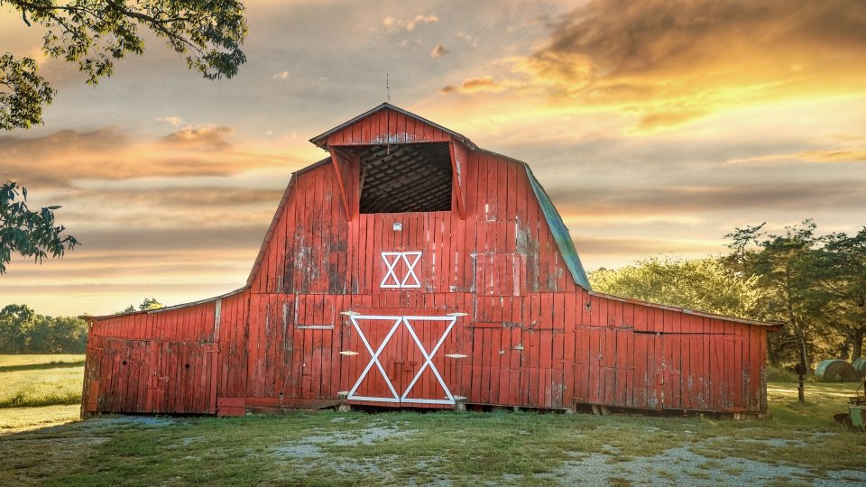 Red barn with distinctive roof against a dramatic sunset sky, surrounded by trees and grass. The scene conveys a rustic, tranquil countryside vibe.