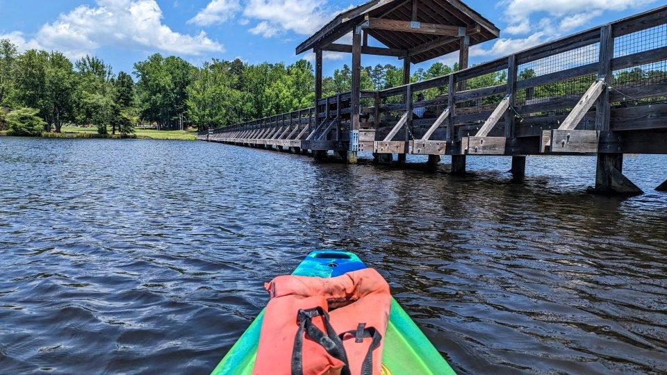 Green kayak with an orange life jacket on a lake near a wooden bridge under a blue sky.