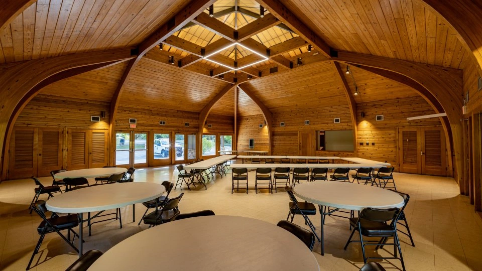 Wooden hall with exposed beams, round tables, and chairs, warmly lit.