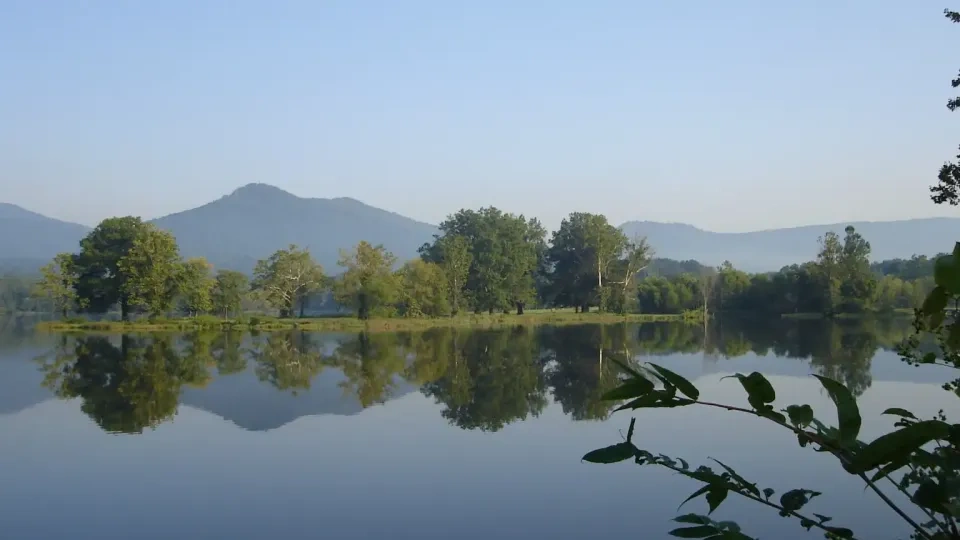Calm lake reflecting trees and distant mountains under a clear blue sky.