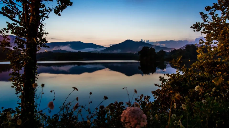 Sunset over a calm lake, mountains and trees reflected in water.