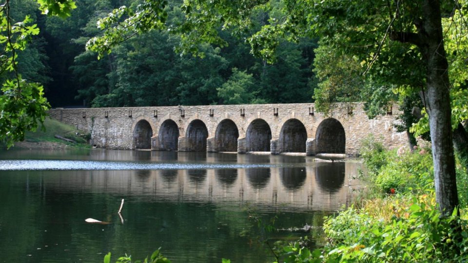 The same long arched stone bridge spans a calm lake, surrounded by dense green summer foliage.