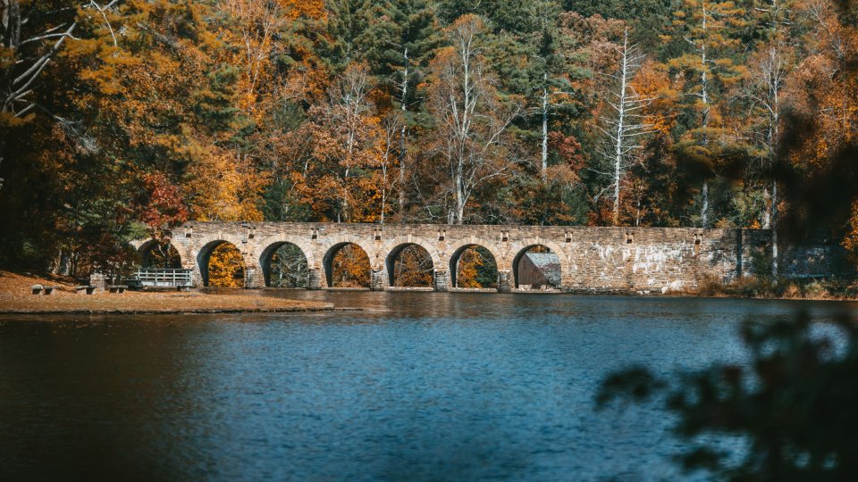 A multi-arched stone bridge spans a lake, surrounded by dense forest with vibrant fall foliage in shades of orange, yellow, and green.