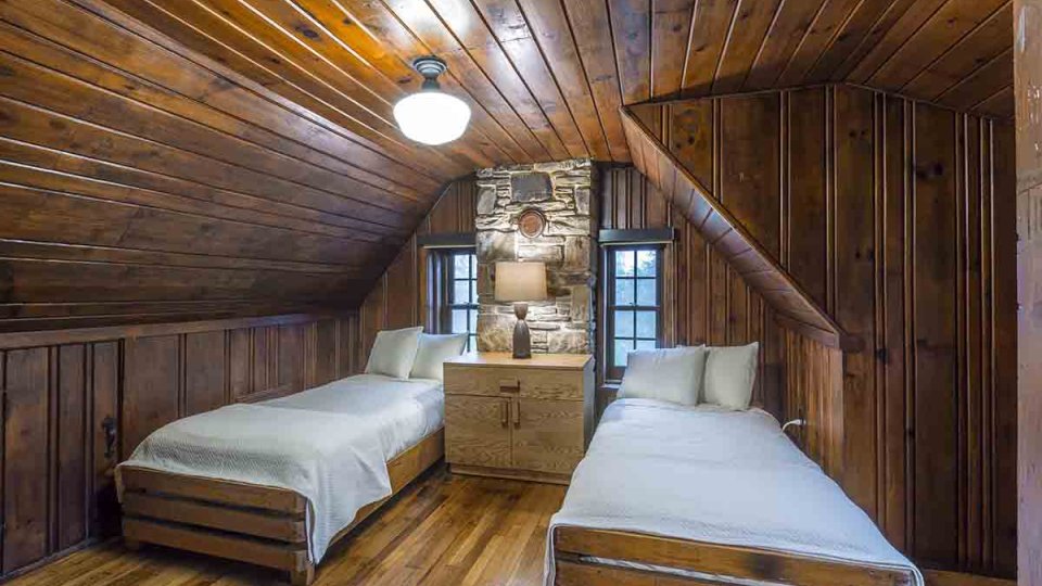 Wooden bedroom with two beds, overhead lighting, and a nightstand dresser with a lamp on top