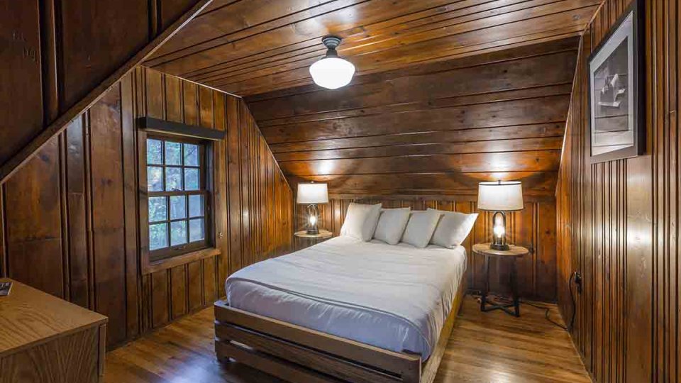 Wooden bedroom with one bed, a dresser, overhead lighting, a window, and a lamp on each side of the bed