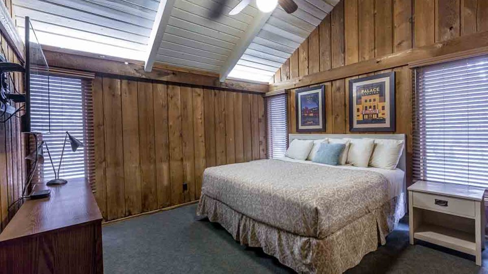 Wooden bedroom with single bed, nightstand, windows, picture frames above bed, fan, dresser, lamp, and mounted television