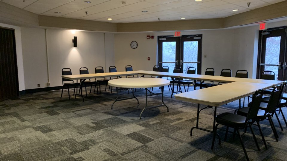 A meeting room with U-shaped tables and chairs arranged around the perimeter, carpeted flooring, and double doors leading outside.