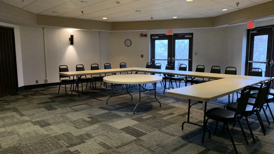 Meeting space with U-shaped tables and chairs, carpeted floor, and natural light from multiple glass doors.