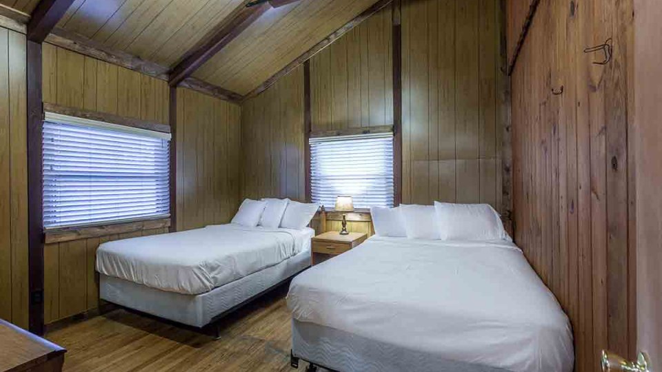 Wooden bedroom with a dresser, two windows, two beds, and a nightstand with a lamp on top in between each bed