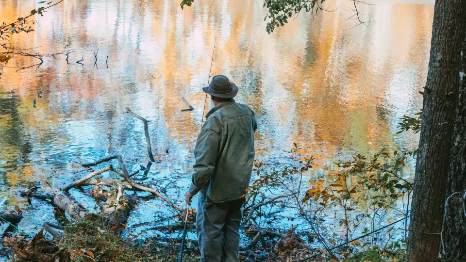 Person wearing a green jacket and wide-brim hat stands on the grassy, wooded edge of a lake, holding a fishing rod as autumn colors reflect on the water.
