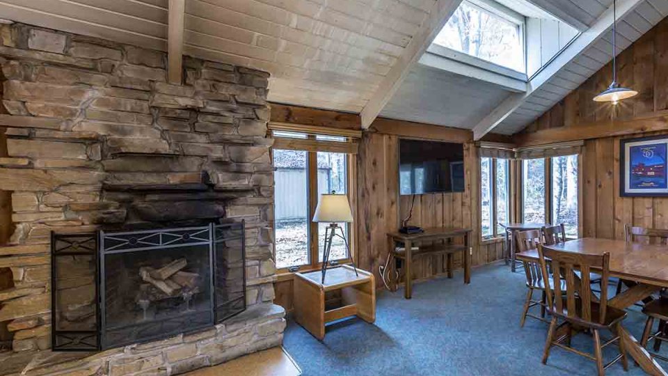 Wooden living room with blue carpet, stone fireplace, mounted television, windows, lamp, dining table, and over head lighting
