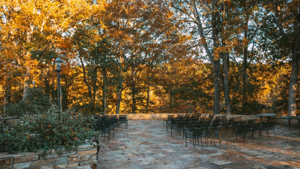 A stone patio with rows of black chairs overlooks a backdrop of tall trees in peak autumn color.
