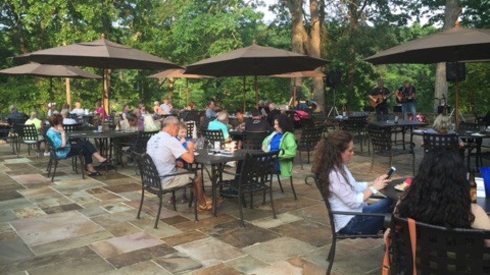 Guests seated under large umbrellas on a stone patio surrounded by trees, enjoying live music and outdoor dining.