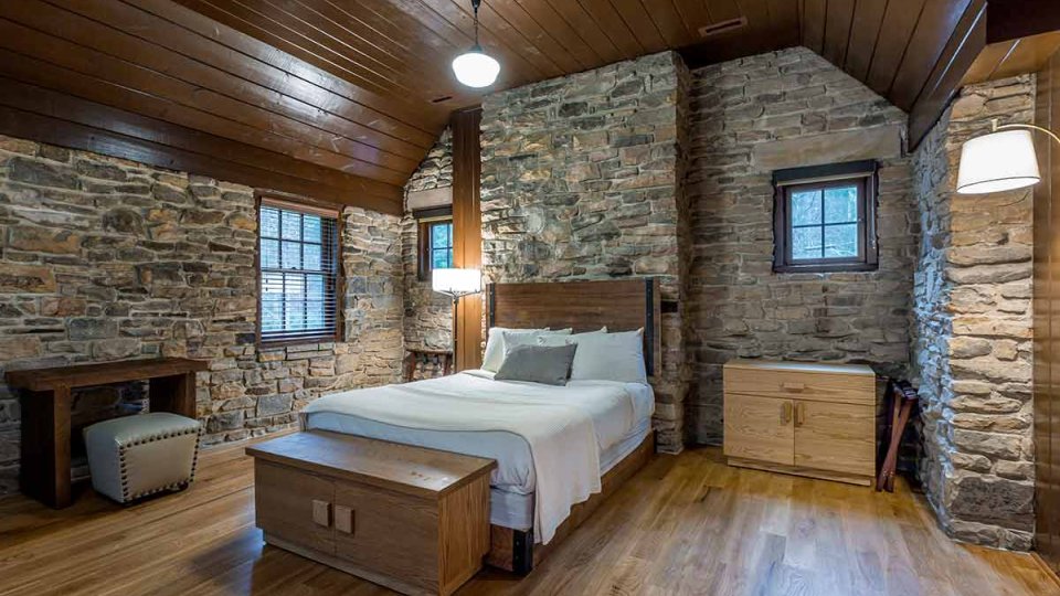 Stone bedroom with one bed, wooden floor and ceiling, cabinets, desk, windows, lamps, and overhead lighting