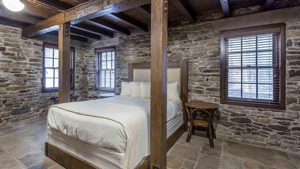 Stone bedroom with one bed, stone floor, wood ceiling, wooden pillars around bed, windows, nightstand, and overhead lighting