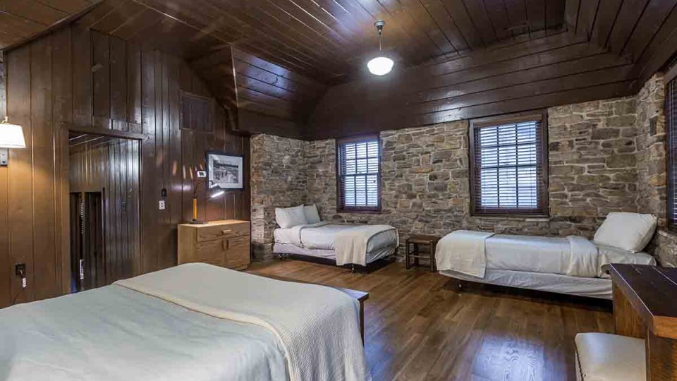 Stone bedroom with three beds, wood floor, wood ceiling, windows, lamps, night stands, and overhead lighting