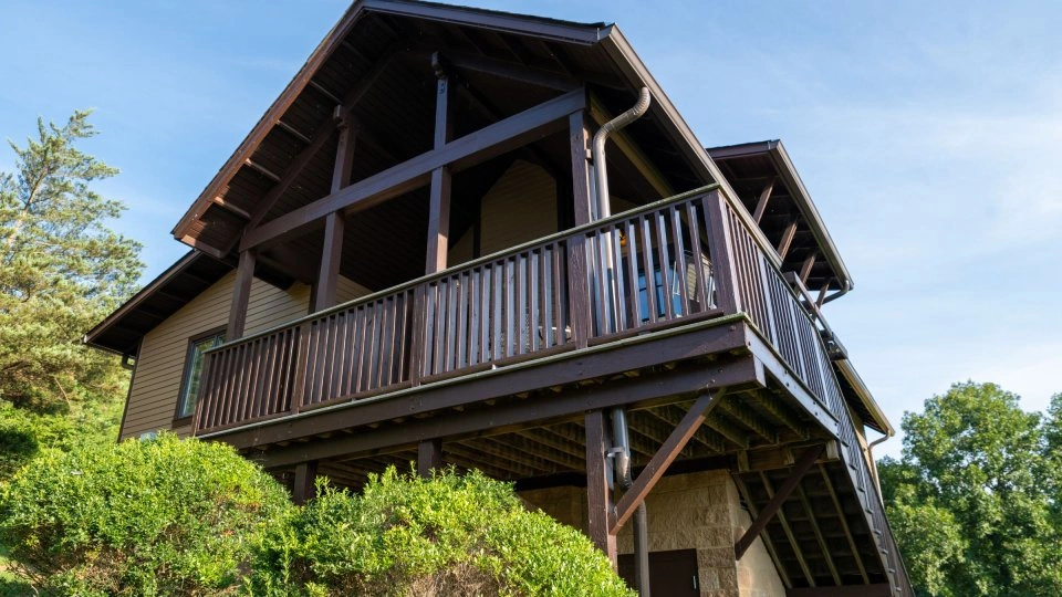 Wooden cabin with a porch, surrounded by greenery, against a blue sky.
