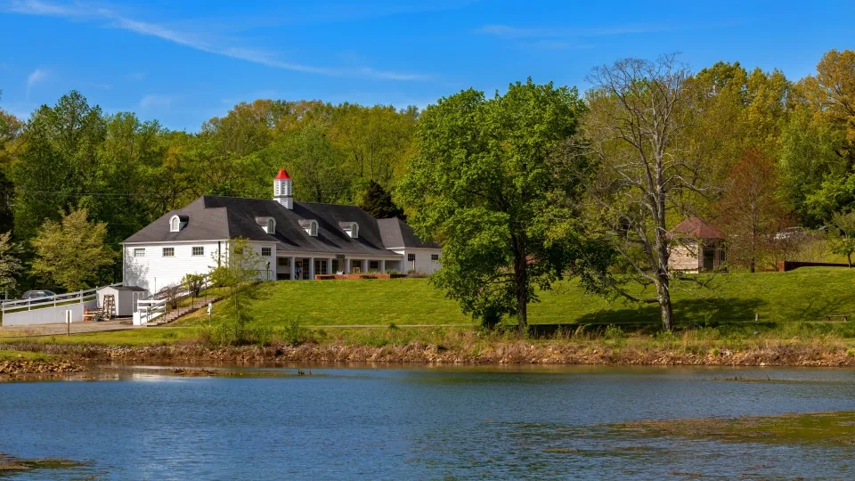 White house with red roof by a pond and trees, under a clear blue sky.