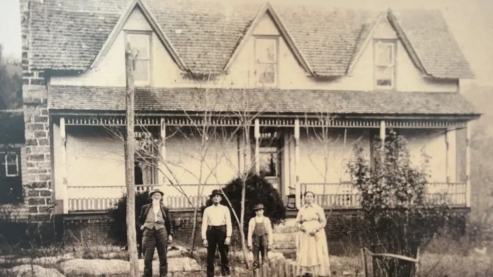 Four people stand in front of a vintage house with a porch and dormer windows.