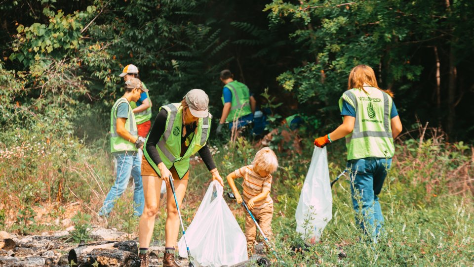 A group of volunteers, including a child, clean a wooded area. They're wearing green vests and using trash bags, conveying teamwork and environmental care.