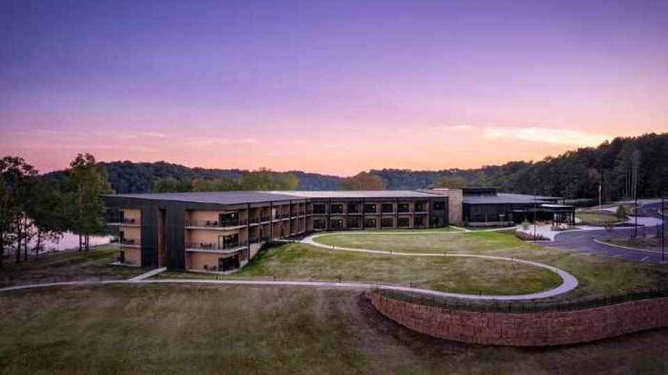Modern lakeside hotel at sunset with a curved design, surrounded by lush greenery and woodland. The sky is a gradient of purple and orange hues.