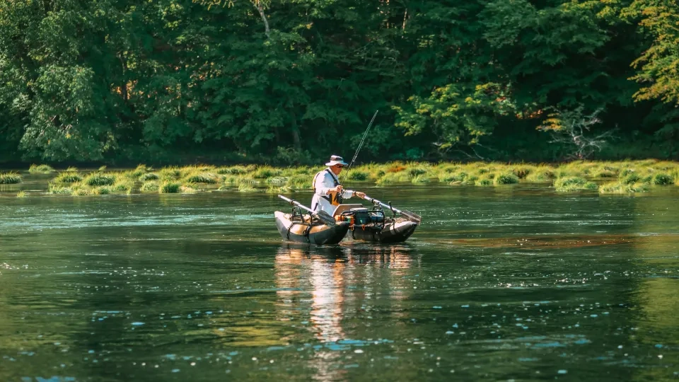 Person fishing from inflatable boat on a calm river, surrounded by lush trees.