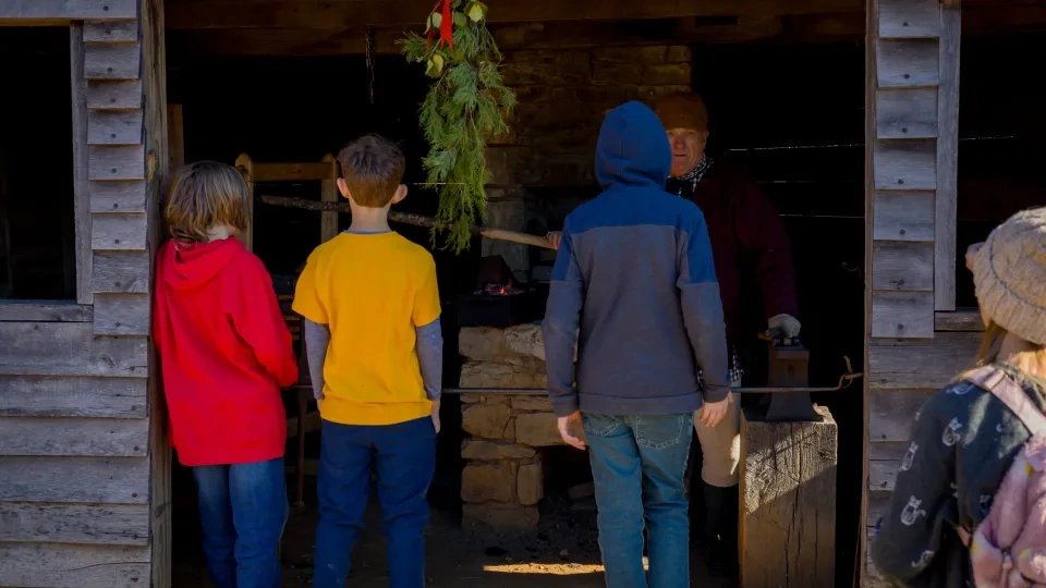 Children in colorful clothes stand in front of a rustic wooden cabin, engaging with an older person inside. A festive wreath hangs above the doorway.